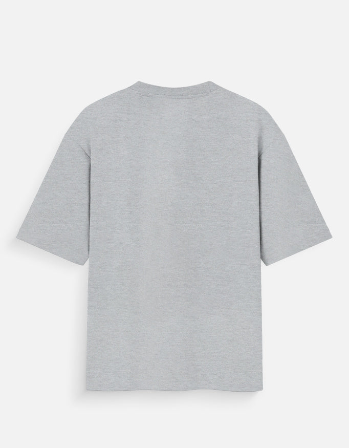 Heather grey oversized unisex t-shirt featuring a centered, small graphic of the word "Aalasya" (laziness) written in red, stylized text with a red strikethrough line, signifying "No Laziness." Soft cotton, drop-shoulder, boxy casual apparel with ironic or statement typography.