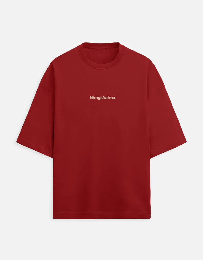 Deep red or maroon oversized unisex t-shirt featuring a centered, small graphic of the text "Nirogi Aatma" (Healthy Soul) written in light brown or gold lettering. Soft cotton, drop-shoulder, boxy casual apparel with a spiritual or motivational typographic design.