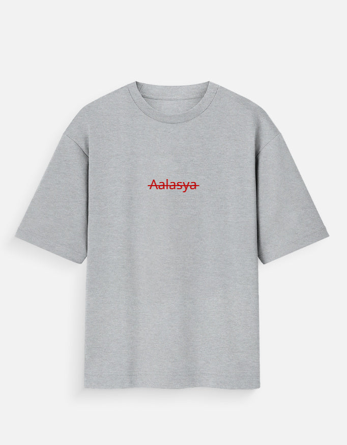 Heather grey oversized unisex t-shirt featuring a centered, small graphic of the word "Aalasya" (laziness) written in red, stylized text with a red strikethrough line, signifying "No Laziness." Soft cotton, drop-shoulder, boxy casual apparel with ironic or statement typography.